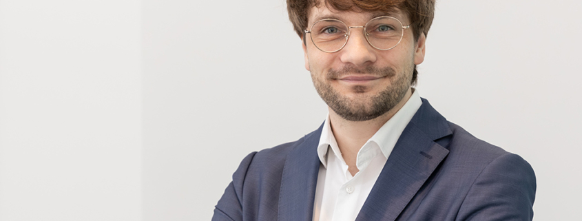 Porträtfoto von Dr. Adrian Steffens, auf dem er mit verschränkten Armen in einem dunkelblauen Jacket und weißem Hemd entspannt in die Kamera lächelt