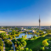 Foto des grünen Münchens aus der Luft, mit Blick auf dem Olympiaturm, das Olympiastadium und den sommerlichen Olympiapark.