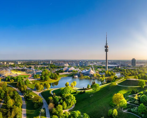 Foto des grünen Münchens aus der Luft, mit Blick auf dem Olympiaturm, das Olympiastadium und den sommerlichen Olympiapark.