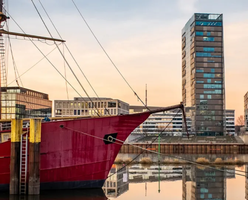 Historisches rotes Schiff im Bremer Hafen vor moderner Stadtkulisse am Wasser als Key Visual für das BOEHMERT & BOEHMERT IP-Seminar in Bremen