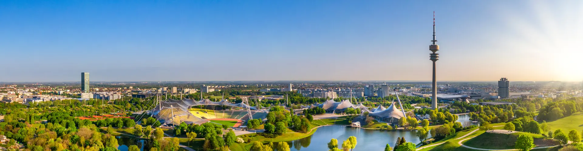 IP-Seminar in München, BOEHMERT & BOEHMERT Schmal zugeschnittenes Panoramafoto von München aus der Luft, mit Blick auf dem Olympiaturm, das Olympiastadium und den sommerlich grünen Olympiapark.